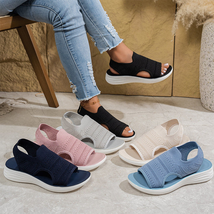 Black sandal worn by a person with multiple color options displayed on a floor.